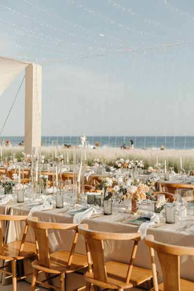 Outdoor wedding reception set up with wooden chairs, a long table decorated with a white table cloth, and peach and pink flowers. The ocean is in the background