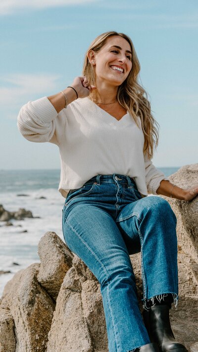 Brand photo of woman sitting on a rock at the beach smiling off camera in a white sweater