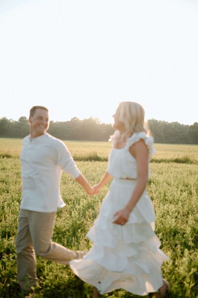 In a romantic scene, a couple dressed in light attire runs hand in hand through a sunlit field, exchanging affectionate glances.