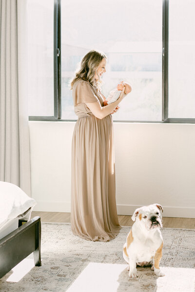 Phoenix mom holding newborn baby facing her while standing in front of window for in-home session. At her feet is an English Bulldog looking at camera.