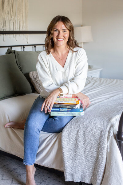 Board certified lactation consultant, Sarah, sitting on a bed wearing a white blouse and jeans holding a stack of pregnancy and lactation books