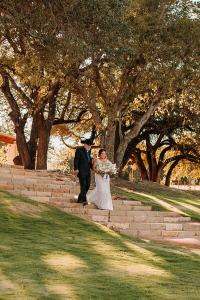 Bride with bouquet and father with cowboy hat walk down steps with grass and trees around them