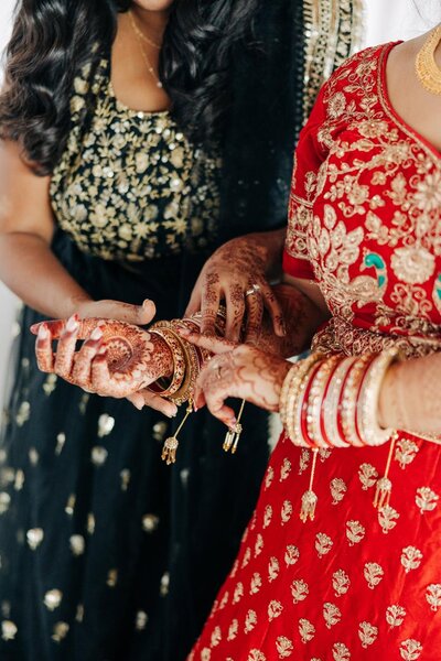 Two individuals dressed in traditional indian attire with one showcasing henna-tattooed hands and both wearing bangles.