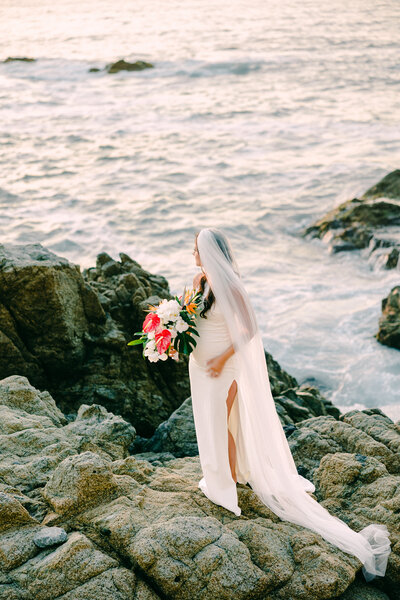 Bride standing on rocky shore with bouquet, ocean waves in background, captured by a Luxury Wedding Photographer.