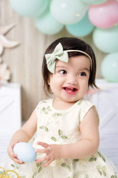 1 year old baby girl holding an egg wearing a green outfit and green ribbon headband