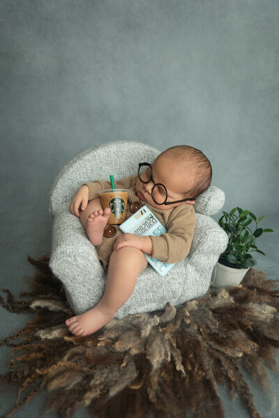 Newborn baby boy alseep on a small couch holding a calcultor, starbucks iced dink & wearing glasses.