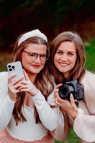 photo of two brunette girls, one is holding an iphone and the other is holding a nikon camera