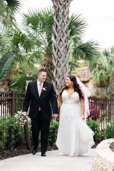 Bride and groom smile at each other as they walk in front of a palm tree