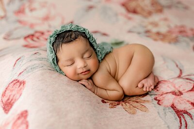 newborn baby girl is curled up on her belly on a flower background and wearing a teal bonnet