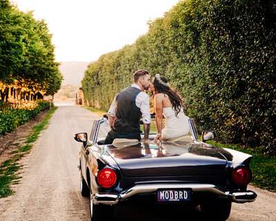 a bride and groom sitting on the back of  a convertible car as they kiss each other
