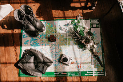 Flat lay of a map, camera, flowers, boots, and wedding details