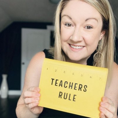 A teacher shows a yellow notebook that says  Teachers Rule with a picture of a ruler.