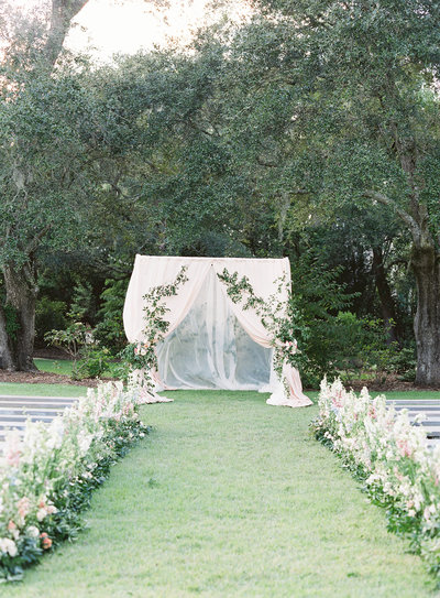 Wedding Ceremony Inspirtation Blush Draped Arbor with Climbing Greenery and Floral Lined Aisle at RiverOaks Charleston