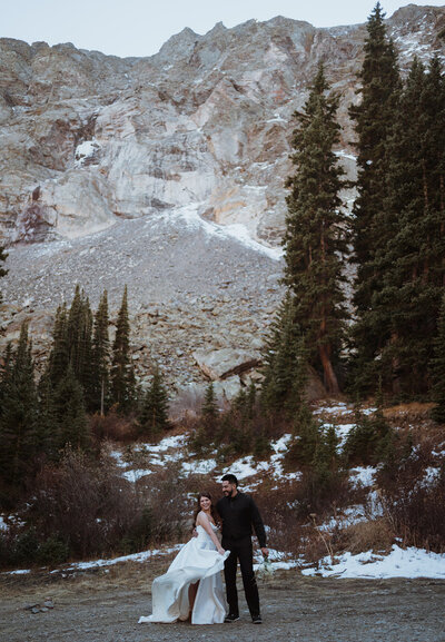 bride and groom are hiking with mountain views as the backdrop. It is summer, and they off roaded in the mountains after saying their vows at an alpine lake with family and friends. The bride is holding her dress as she stands next to the groom in a black suit.