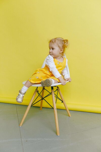 A young child in a yellow dress sits on a wooden chair against a matching yellow background, looking to the side.