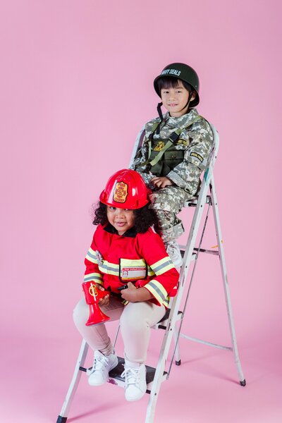 Two children dressed in costumes are sitting on a stepladder against a pink background: one is wearing a U.S. Navy SEAL uniform with a helmet, and the other is dressed as a firefighter in a red uniform with a fire helmet, holding a toy megaphone and fire extinguisher.