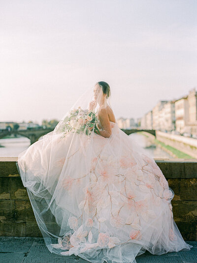 bride sitting on the edge of a bridge in Florence Italy with her veil draped over her head and the city in the background