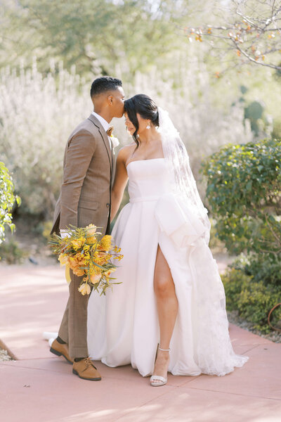 southern california wedding photography of couple cuddling on a manicured path. Bride is wearing designer wedding gown and groom is wearing custom tan suit