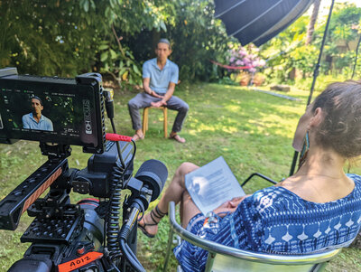 A man being interviewed sits in a chair on grass in front a camera equipment. The interviewer sits next to the camera in the foreground.