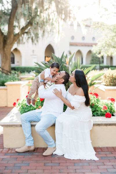 A family of three sitting down in front of flowers while the dad kisses his son. 