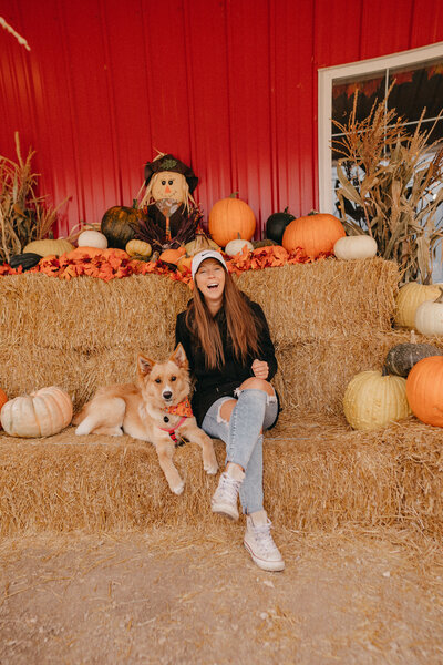 woman laughing with her dog beside her in a pumpkin patch