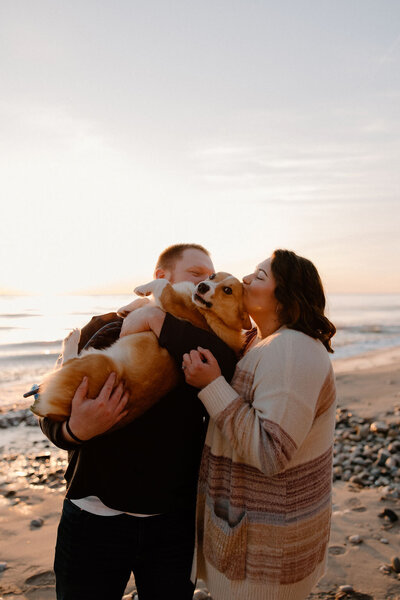 Sweet engagement photoshoot of a couple on the beach at sunset with the man holding their dog and both the man and woman kissing the dog on the head captured by Morgan Ashley Lynn Photography in Milwaukee, WI on the beach of Lake Michigan.