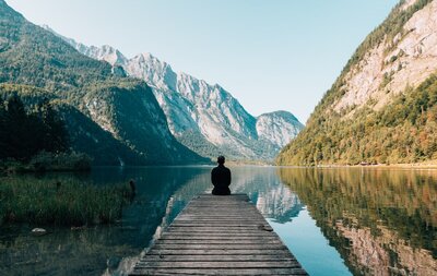 person sitting on a dock in a lake looking at mountains