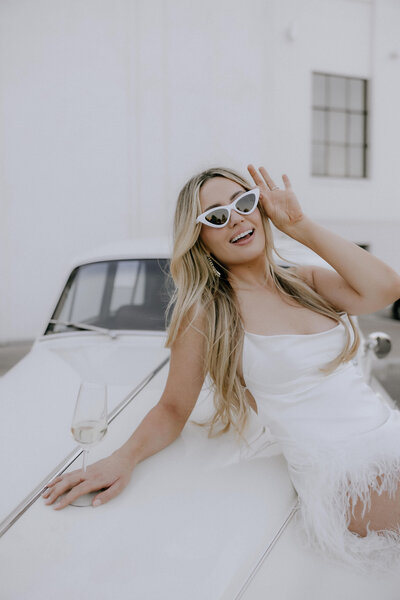 bride holding white glasses leaning on a vintage car