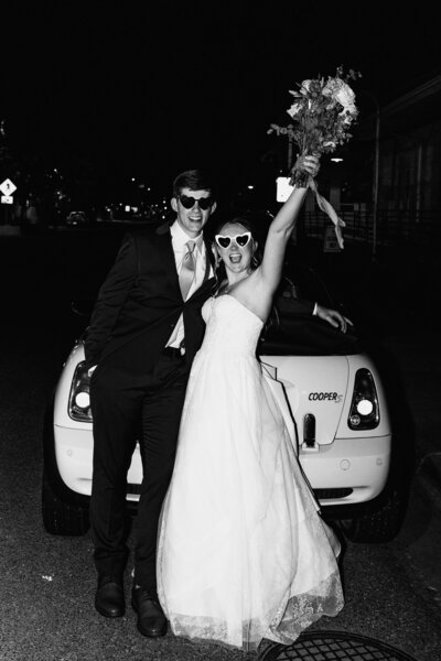 Excited couple poses in front of car wearing heart-shaped sunglasses.
