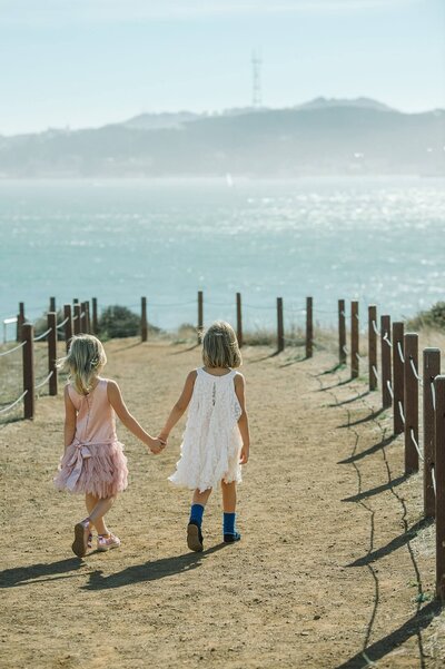 Girl twins in dresses back to camera walking hand in hand going towards the water in Sausalito