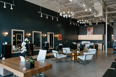 Interior photo of SCENE Salon reception and waiting area. Dark moody walls, concrete floors, and white leather modern chairs, with gold accents and colorful artwork.