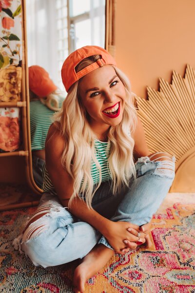 Founder of Infinite Productions, Andi Sweeny, sitting cross legged on a floor wearing ripped jeans, a blue and white striped shirt, and an orange hat.