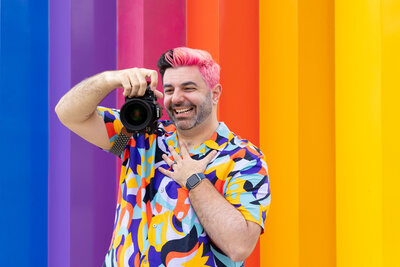 A photographer holding their camera up and laughing in front of a rainbow background.