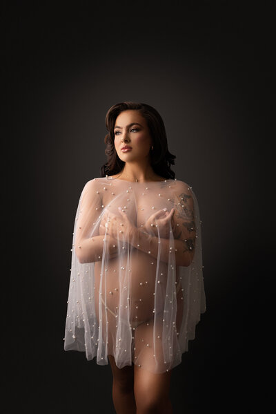 Woman poses for fine art maternity photos in Philadelphia.  She is sitting on her knees angled away from the camera. She is wearing an off-the-shoulder dusty rose gown with her cleavage exposed. The shoulder of the gown drapes along side of her and the gown is puddled around her. The woman is gently holding her bump. The woman's eyes are closed and slightly angled up toward the light. Captured by best Philadelphia maternity photographer Katie Marshall.