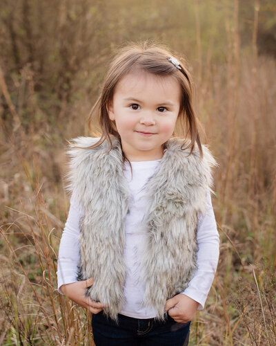 little girl with brown hair and big brown eyes in fur vest standing in a field of tall grass
