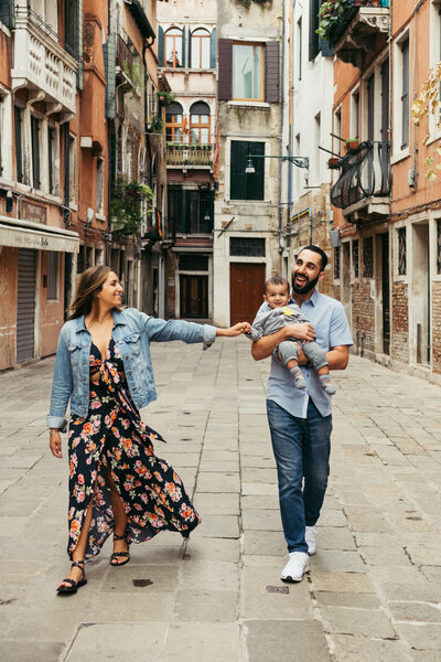 This heartwarming photo captures a couple and their toddler strolling through the picturesque alleys of Venice, beaming with smiles and joy. The father lovingly carries the baby in his arms, while the family takes in the breathtaking scenery of the city. A perfect moment frozen in time, showcasing the beauty and charm of Venice as well as the love and happiness of this family
