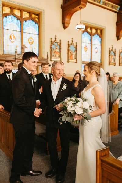 Bride smiles at groom as her dad hands her off at the beginning of their wedding ceremony at St Anthony of Padua Catholic Church