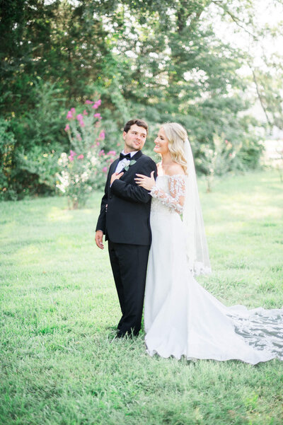 groom in black suit and bride in white gown look at one another in a green field