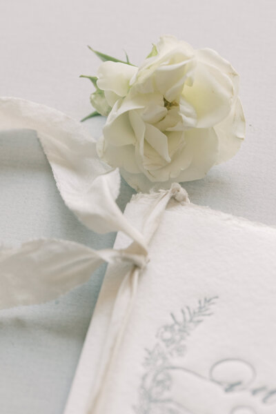 White flower and part of a wedding invite with cream ribbon on a soft pale blue background.