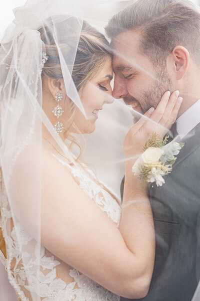 Bride and groom touching forheads under veil with brides hand on grooms face