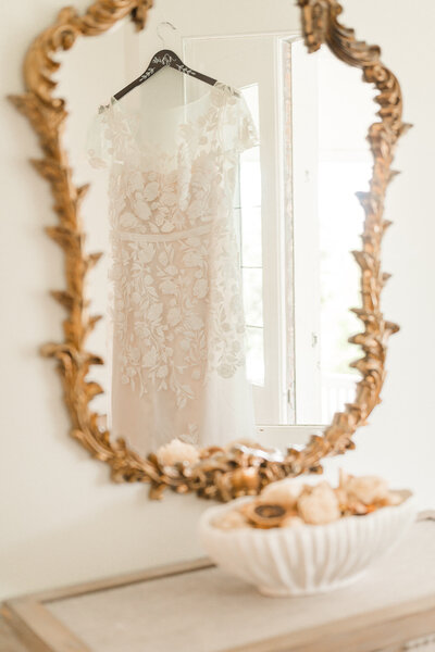 A lace wedding dress hanging on a door and in the reflection of a mirror by JoLynn Photography, a Raleigh wedding photographer