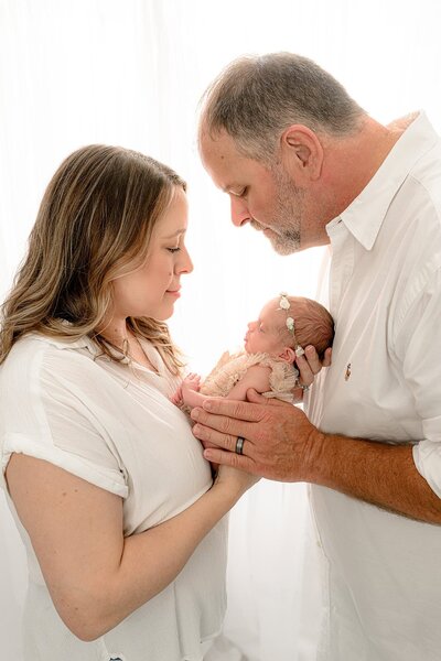 New mom and Dad dressed in white hold their newborn daughter between then 