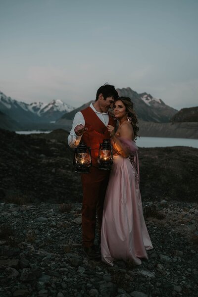 A couple in formal attire holding lanterns stands close together outdoors with mountains in the background at dusk, capturing the spirit of a Utah desert photography retreat focused on real connection and growth.