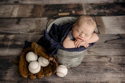 newborn boy is posed in a bucket sitting on a wooden floor with baseballs and glove beside him