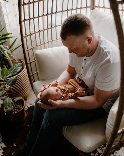 Adorable, dark and vintage looking newborn photography session by Morgan Ashley Lynn Photography in Madison, WI of a dad sitting in a boho inspired rattan couch chair holding his baby boy in his lap and looking down at him with plants in front of them