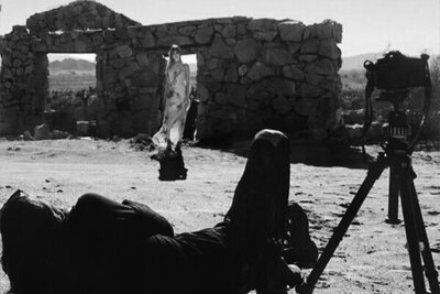 BTS black and white Mark Maryanovich photographing female musician Kendall Rucks standing in front of stone building in desert wearing long dress tripod beside him