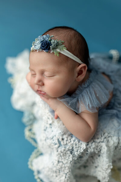 Newborn baby girl in blue posed in a box with white lace and a flower headband.