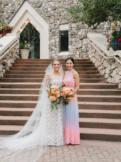 Bride in lace dress, and her bridesmaid smile in front of a grand outdoor staircase in front of a grey stone building.