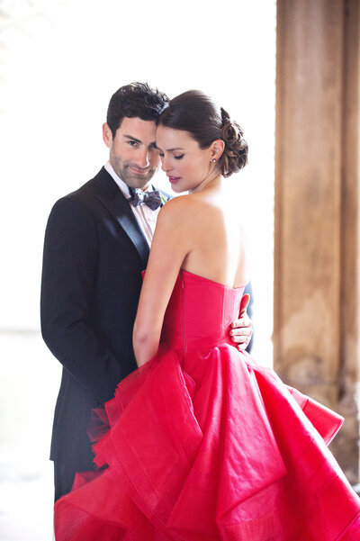 Woman in red ballgown leaning against man in a suit against a stone wall