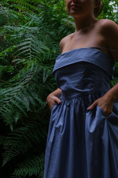girl in a blue dress in front of green bushes with hands in pockets